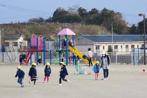 園庭で、黄色い帽子を被った幼児が走り回って遊んでおり、その奥に複合遊具の滑り台が写っている写真