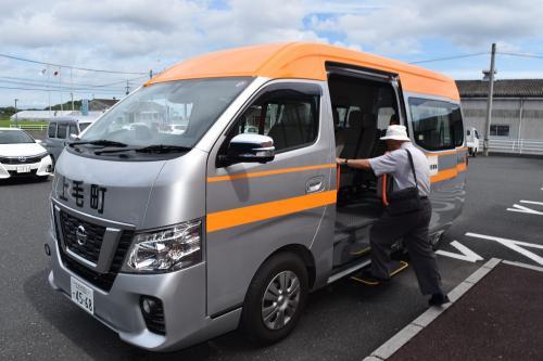 車体がグレーでフロント部分に「上毛町」と書かれており、天井部分とドアはオレンジ色で塗られているコミュニティバスに男性が乗り込もうとしている写真