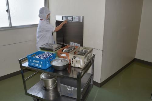 牛乳や食缶の乗った給食ワゴンが給食リフトの前に置かれており、白衣と帽子を身に着けた職員の方が、給食用リフトの操作ボタンを操作している写真