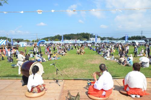 広場手前のステージで太鼓や横笛を演奏している3人の人がおり、広場の芝生には多くの人が集まり、奥には青と白の縞模様のテントが沢山張られている写真