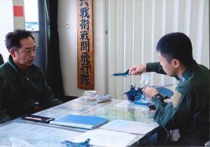 町長と自衛隊員が机に向き合って座り、戦闘機の模型を手に持って話をしている自衛隊員を見ている町長の写真