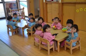 教室に机が2台置かれており、青いスモッグを着た4名の幼児と、赤いスモッグと青いスモッグを着た幼児6名がそれぞれの机を囲んで椅子に座って、カメラ目線で写っている写真