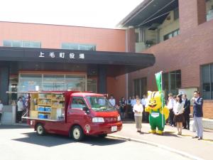 上毛町役場前に止っている商品を乗せた赤い移動販売車、グリーンコープのマスコットキャラクターと移動販売車の後ろに集まった人たちが拍手をしている写真